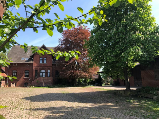 Hof Holthöfer Ein rotes Backsteingebäude auf einem Steinhof, umgeben von grünen und roten Laubbäumen.