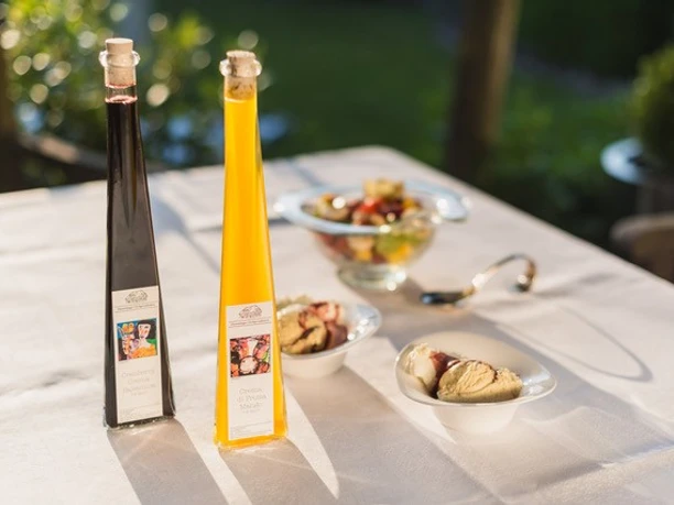 Hümmlinger Hofspezialitäten.jpg Zwei schlanke Flaschen mit Likören auf weißer Tafel, daneben Dessertschalen im Sonnenlicht draußen
