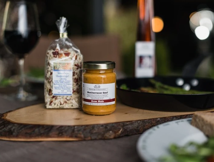 Hümmlinger Hofspezialitäten_3.jpg Glas Honig und regionale Feinkost auf Holzbrett mit Wein im Hintergrund in stimmungsvoller Atmosphäre