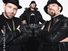 Drei Männer in schwarzer Lederkleidung und Hüten posieren selbstbewusst vor einem DJ-Pult.