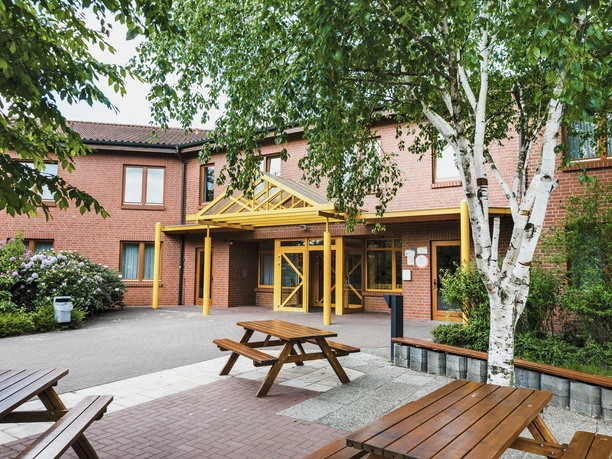 Lingen_20150521_0172.jpg Backsteingebäude mit gelbem Eingangsbereich, Sitzbänken im Hof und umgeben von Bäumen.