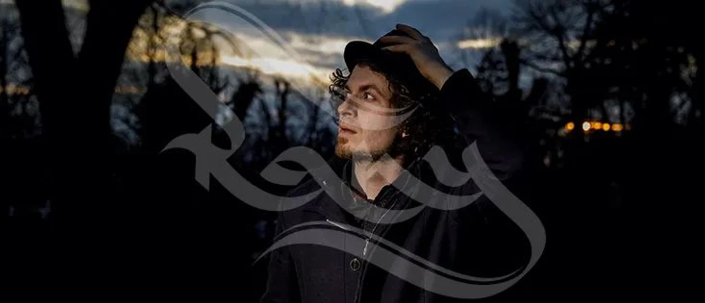 Ein Mann mit lockigem Haar und Hut steht im Dunkeln, sein Gesicht wird von einer Lichtquelle beleuchtet.