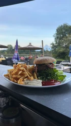 Heimser Hof Ein Teller mit Burger und Pommes frites auf einer Terrasse vor grüner Wiese und Sonnenschirmen.