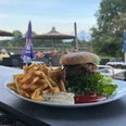Heimser Hof Ein Teller mit Burger und Pommes frites auf einer Terrasse vor grüner Wiese und Sonnenschirmen.