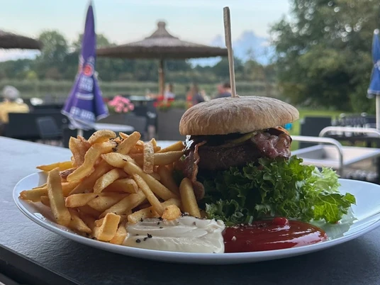 Heimser Hof Ein Teller mit Burger und Pommes frites auf einer Terrasse vor grüner Wiese und Sonnenschirmen.
