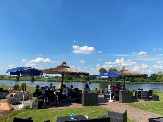 Heimser Hof Besucher sitzen unter Strohdächern an Tischen am Ufer eines ruhigen Sees unter blauem Himmel.