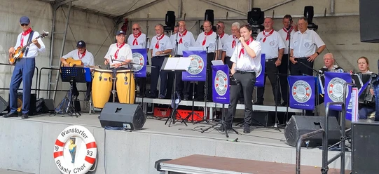 Wunstorfer-Shantychor-2025.jpg Ein Chor in maritimer Kleidung singt auf einer Bühne, begleitet von Instrumenten wie Gitarre und Congas.