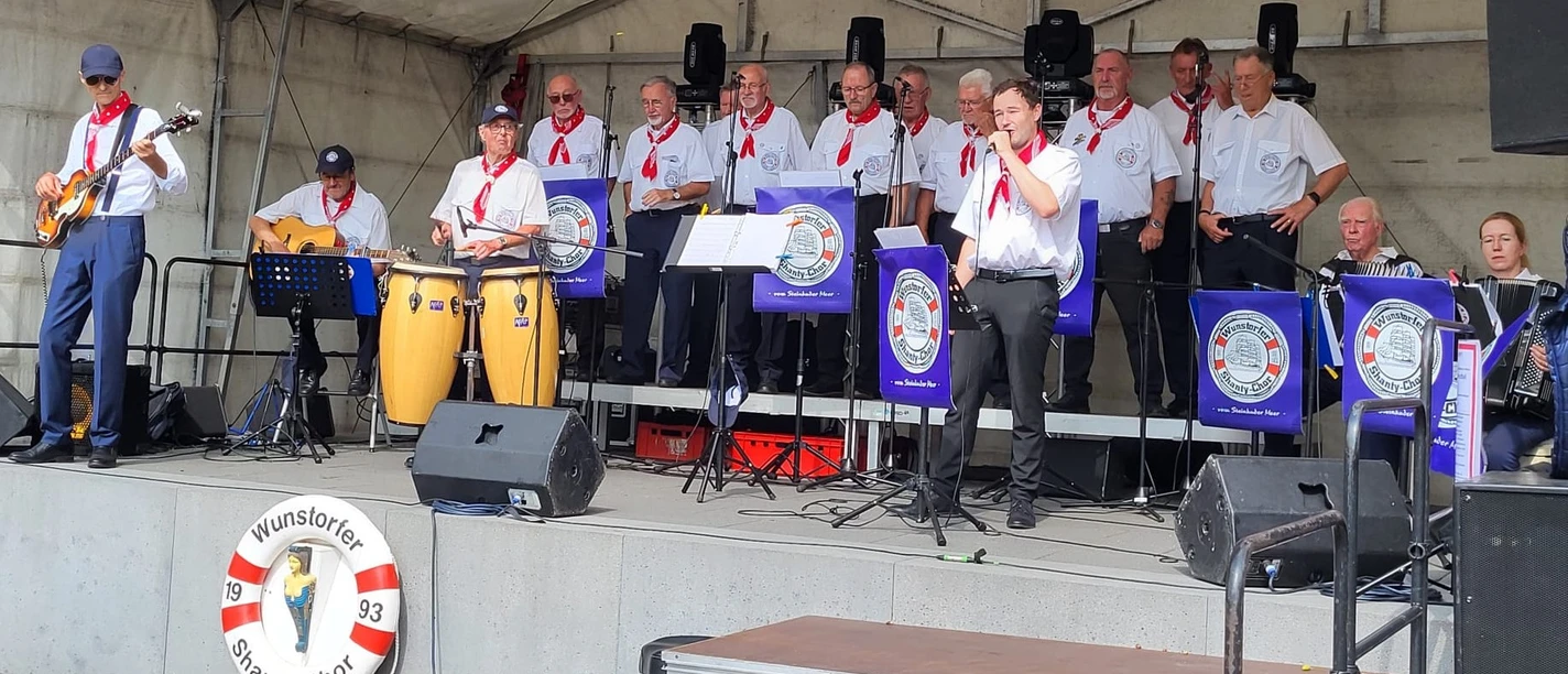 Wunstorfer-Shantychor-2025.jpg Ein Chor in maritimer Kleidung singt auf einer Bühne, begleitet von Instrumenten wie Gitarre und Congas.