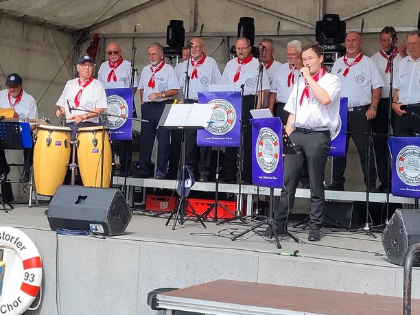 Wunstorfer-Shantychor-2025.jpg Ein Chor in maritimer Kleidung singt auf einer Bühne, begleitet von Instrumenten wie Gitarre und Congas.