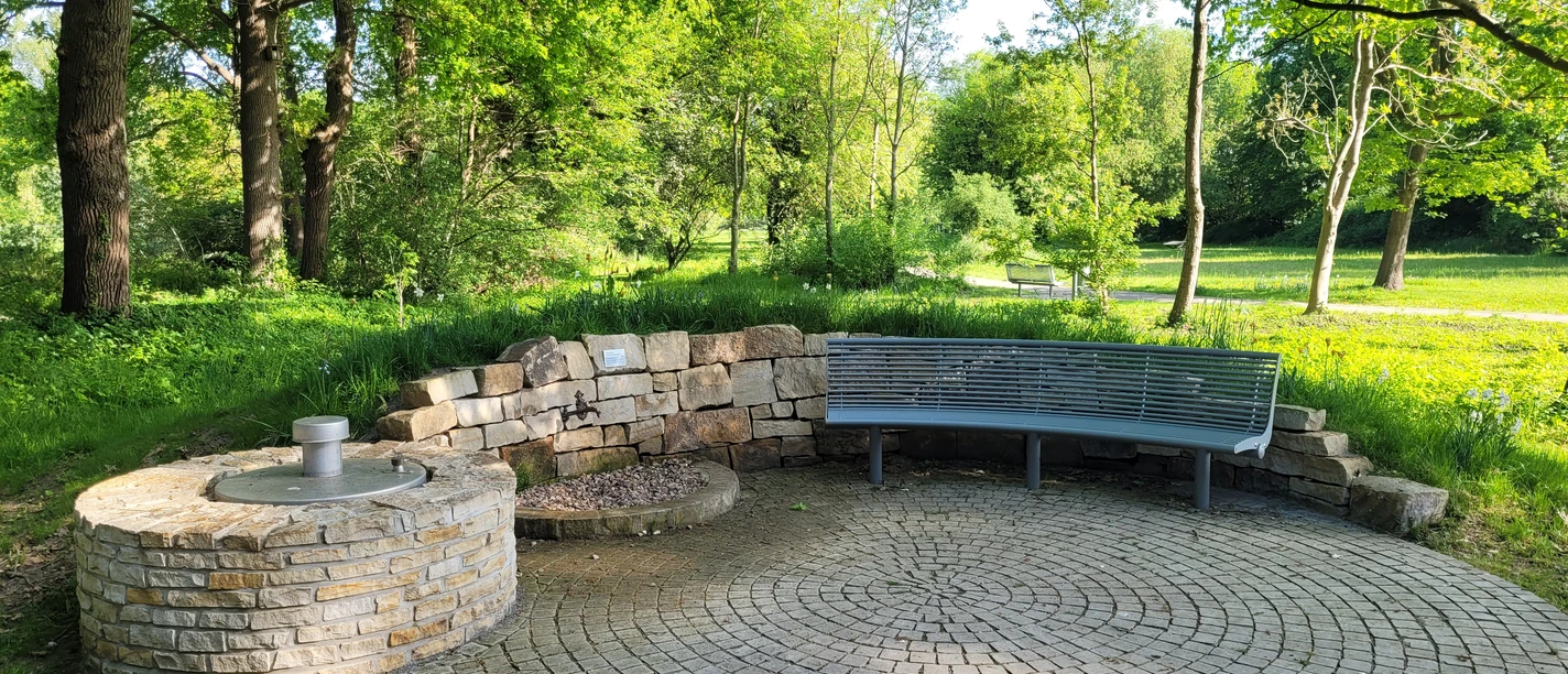 Kurpark Levern Runder Platz im Kurpark Levern mit Sitzbank und Brunneneinfassung, umgeben von grünen Bäumen.