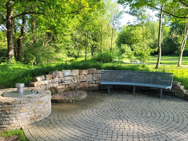 Kurpark Levern Runder Platz im Kurpark Levern mit Sitzbank und Brunneneinfassung, umgeben von grünen Bäumen.