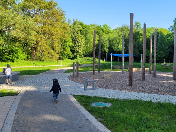 Kinder laufen im Kurpark Levern auf kurvigen Wegen, umgeben von hohen Bäumen und Spielgeräten.
