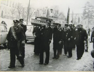 Een groep mannen in traditioneel uniform draagt een groot modelschip tijdens een parade.
