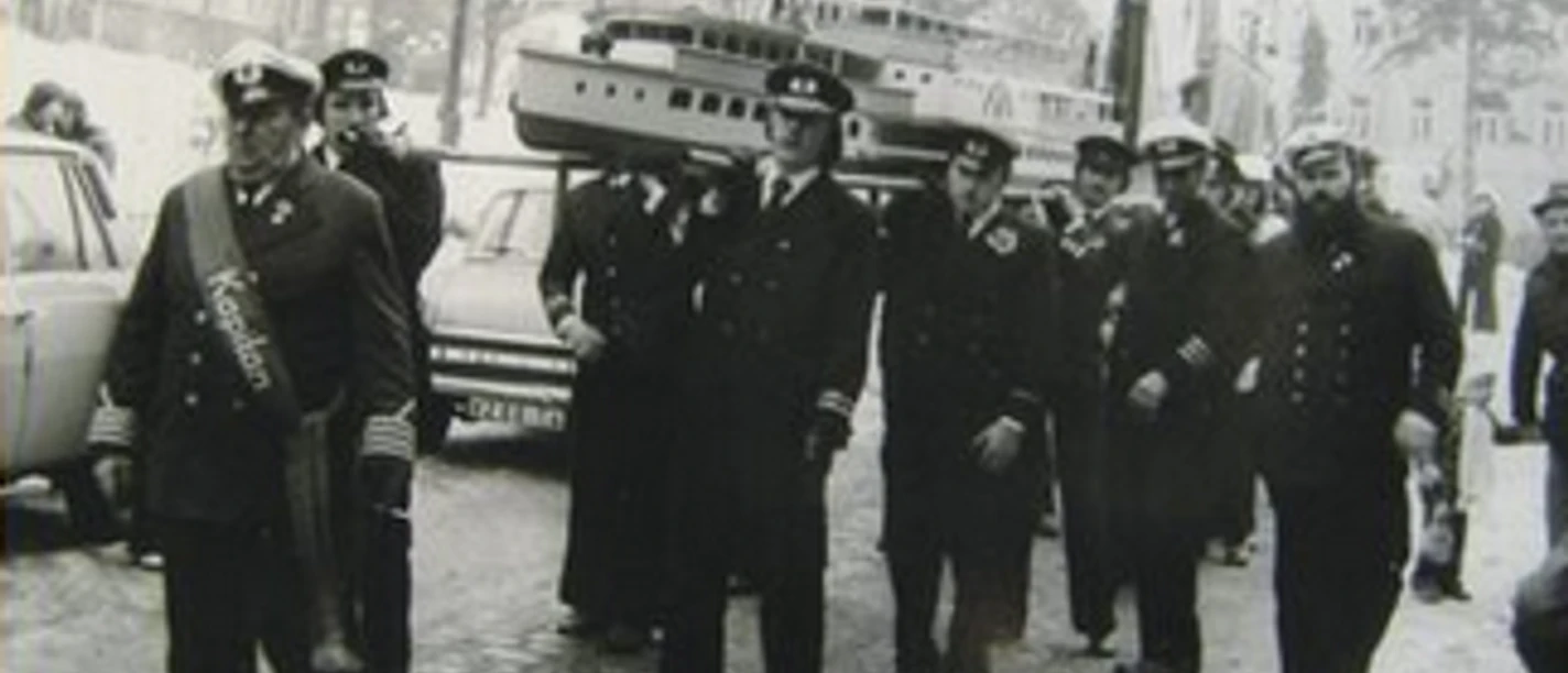 A group of men in traditional uniform carry a large model ship during a procession.