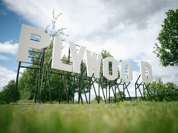 Purple Path_Werk__Plywood__Jay Gard_Foto Johannes Richter.jpg Große Buchstaben "PLYWOOD" auf Metallstruktur, umgeben von grünen Bäumen und blauem Himmel.Large letters "PLYWOOD" on metal structure, surrounded by green trees and blue sky.Velká písmena "PLYWOOD" na kovové konstrukci, obklopená zelenými stromy a modrou oblohou.Duże litery "PLYWOOD" na metalowej konstrukcji, otoczone zielonymi drzewami i błękitnym niebem.Grote letters "PLYWOOD" op metalen structuur, omringd door groene bomen en blauwe lucht.Grandi lettere "PLYWOOD" su una struttura metallica, circondata da alberi verdi e cielo blu.
