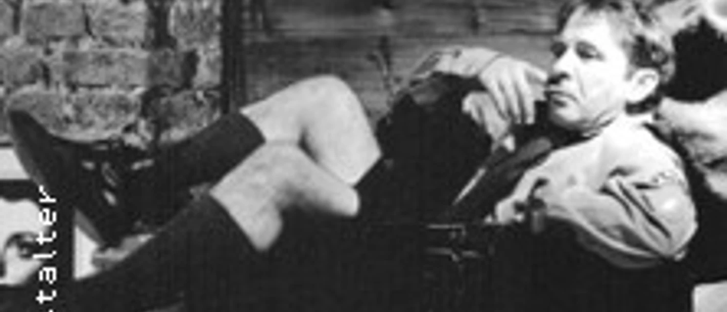 Ein entspannt sitzender Mensch vor einer Backsteinwand, lässig gekleidet in Shorts und Sakko.