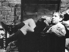 Ein entspannt sitzender Mensch vor einer Backsteinwand, lässig gekleidet in Shorts und Sakko.