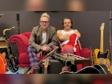 Zwei Musiker in entspannter Pose auf einer roten Couch, umgeben von Gitarren und Saxofonen.