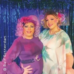 Zwei Drag Queens in farbenfrohen Kostümen und Perücken posieren vor einem glitzernden Hintergrund.