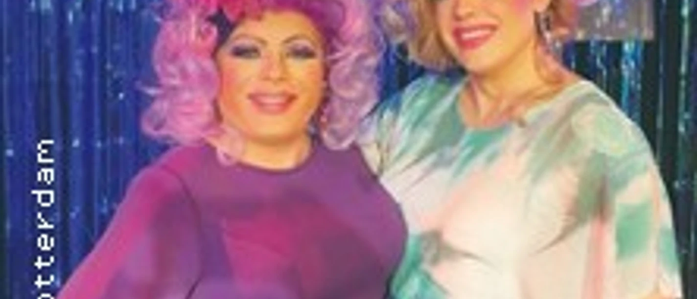 Zwei Drag Queens in farbenfrohen Kostümen und Perücken posieren vor einem glitzernden Hintergrund.