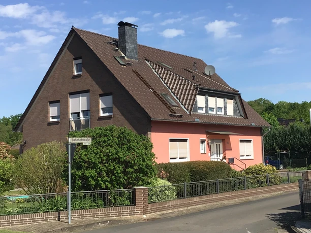 Een huis van twee verdiepingen met een rode gevel en bruine daken, omringd door groene struiken en bomen.