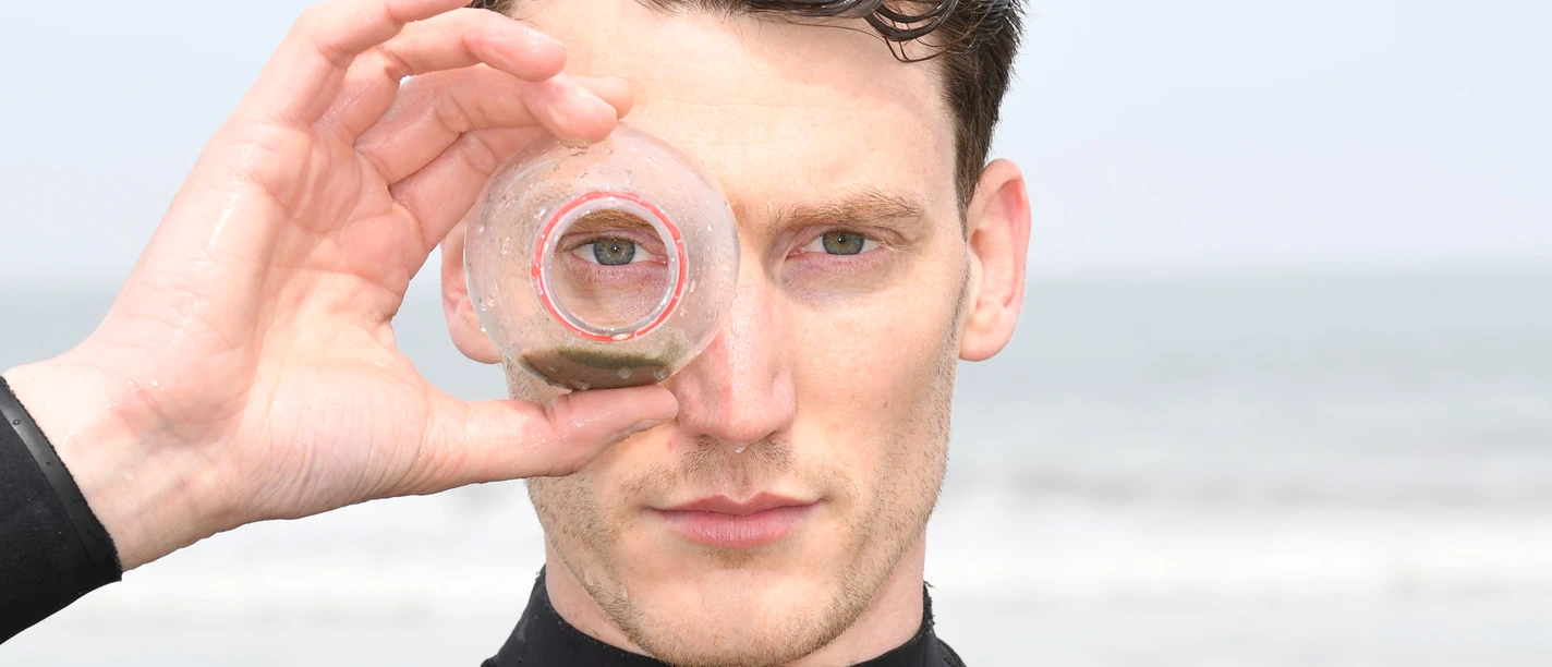 Chris Weigand Chris Weigand im Neoprenanzug am Meer schaut durch eine Plastikflasche in die Kamera.