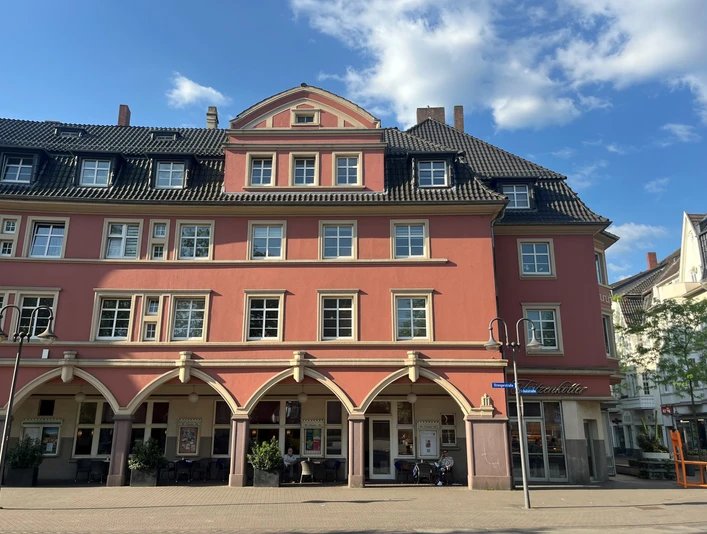 Außenansicht Konditorei & Café Fritzenkötter Ein rot gestrichenes, mehrstöckiges Gebäude mit Arkaden vor blauem Himmel und vorbeiziehenden Wolken.