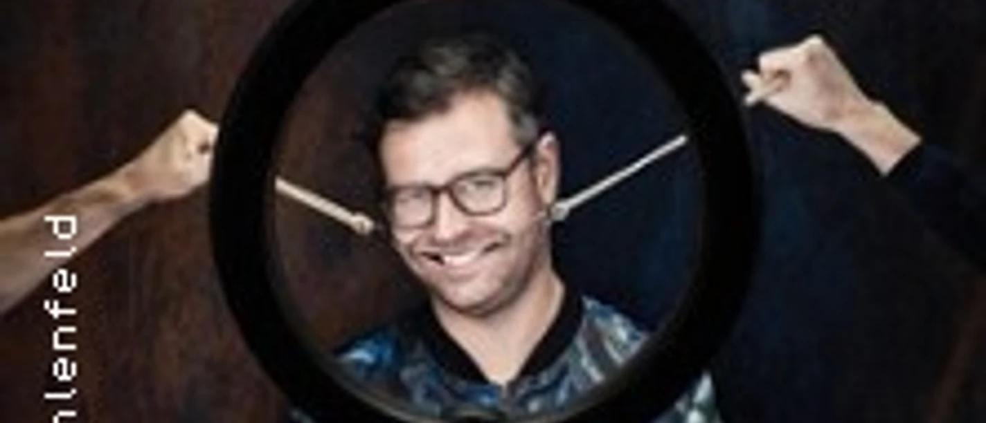 Ein Mann mit Brille lächelt, während er von zwei Händen mit Drumsticks umrahmt wird.