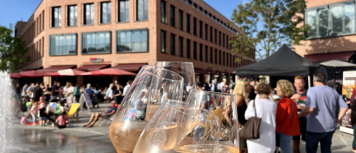 Feierabendmärkte Monheim am Rhein Mehrere Personen stoßen mit Weingläsern an einem belebten Markt auf einem sonnigen Platz an.