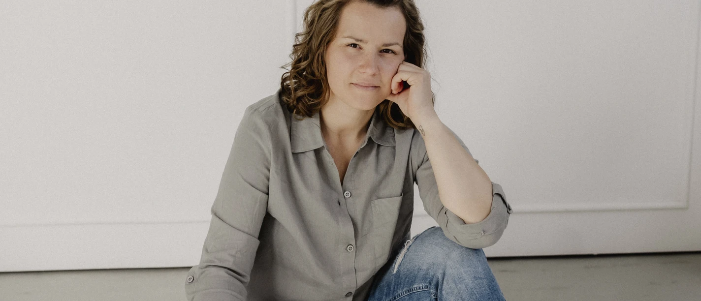 Annette Müller Eine Frau in Jeans und grauer Bluse sitzt barfuß entspannt auf dem Boden in hellem Raum.