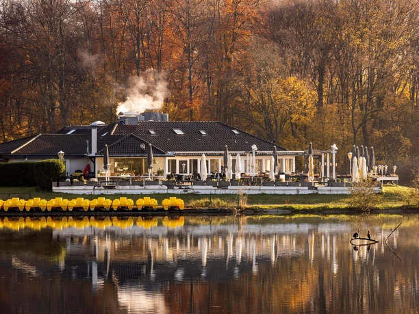 Decksteiner Weiher Ein herbstlich gefärbter Wald umrahmt ein charmantes Gebäude am Decksteiner Weiher, dessen Wasser die Umgebung spiegelt.