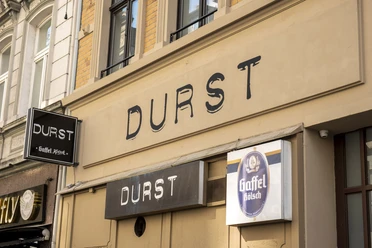 Durst Illuminated sign reading ‘Durst’ (Thirst) on the front of a building with a Kölsch sign on a historic street.