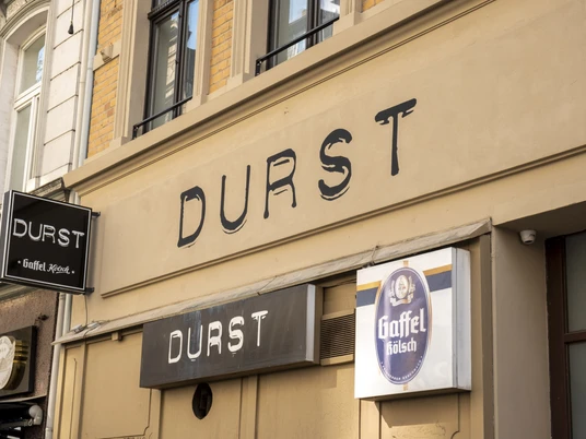 Durst Leuchtreklame „Durst“ auf Gebäudefront mit Gaffel Kölsch-Schild an einer historischen Straße.Illuminated sign reading ‘Durst’ (Thirst) on the front of a building with a Kölsch sign on a historic street.