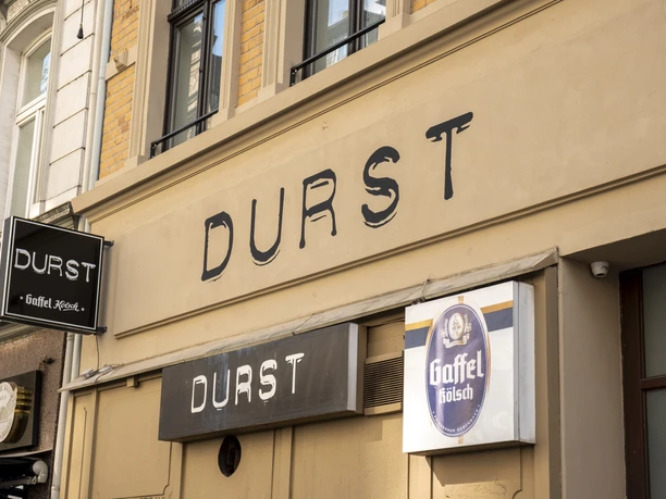 Durst Illuminated sign reading ‘Durst’ (Thirst) on the front of a building with a Kölsch sign on a historic street.
