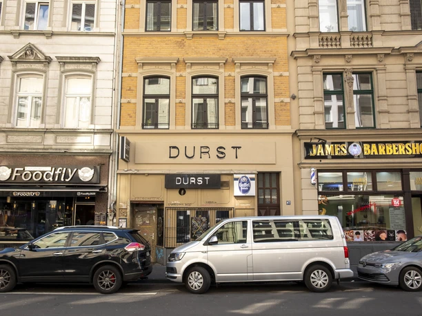 Durst The street scene in Cologne shows the ‘Durst’ building between a hairdressing salon and a restaurant, with parked cars along the street. The word ‘DURST’ dominates the façade in clear, modern lettering.