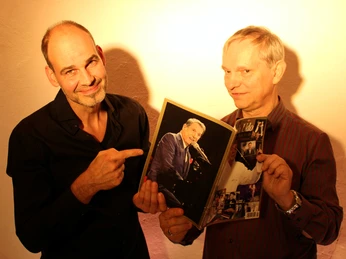 Pressefoto_1_UDO-Programm.jpg Roland und Klaus, zwei Männer mittleren Alters, halten lächelnd ein Bild von Udo Jürgens in die Kamera.