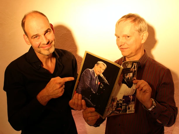 Pressefoto_1_UDO-Programm.jpg Roland und Klaus, zwei Männer mittleren Alters, halten lächelnd ein Bild von Udo Jürgens in die Kamera.