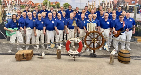 Shantybenthullen.jpg Ein Chor von Männern in blauen Hemden posiert vor einem maritimen Hintergrund mit Musikinstrumenten.