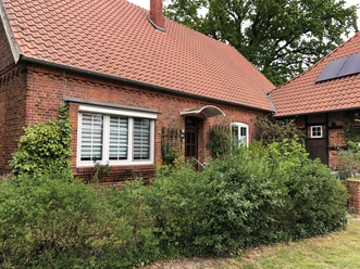 Aller-Rose Ein Backsteinhaus mit Giebeldach und rankenden Pflanzen, umgeben von grünen Sträuchern und Bäumen.
