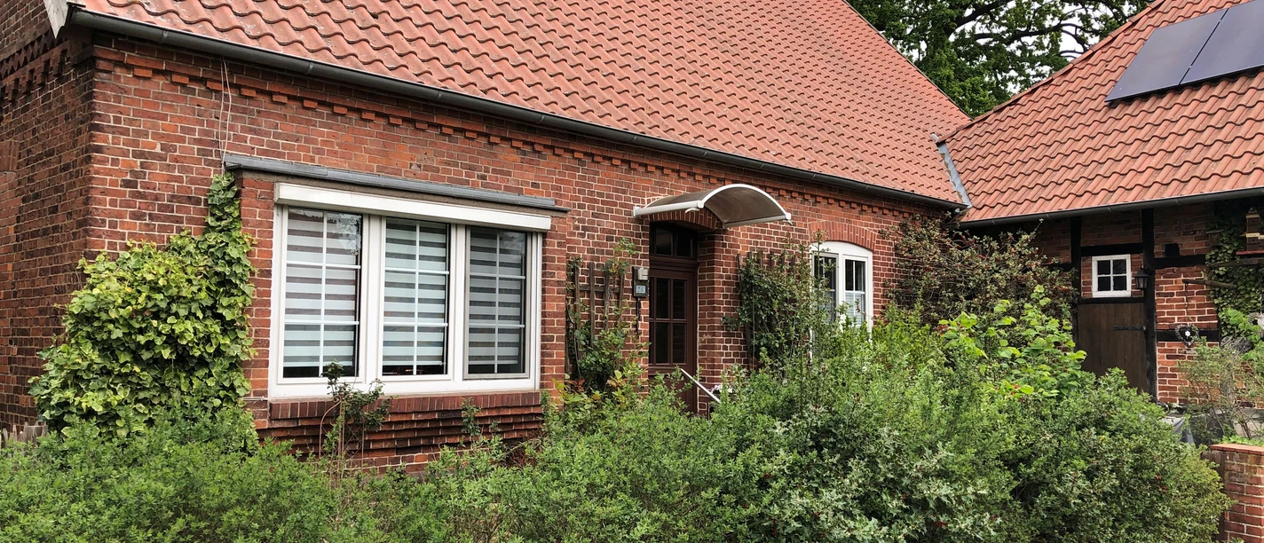 Aller-Rose Ein Backsteinhaus mit Giebeldach und rankenden Pflanzen, umgeben von grünen Sträuchern und Bäumen.