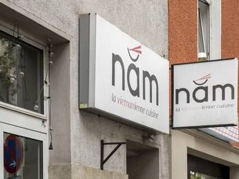 Nam La Vietnamienne Cuisine Fassadenansicht eines vietnamesischen Restaurants namens "nâm" mit markantem Logo.Facade view of a Vietnamese restaurant called ‘nâm’ with a striking logo.