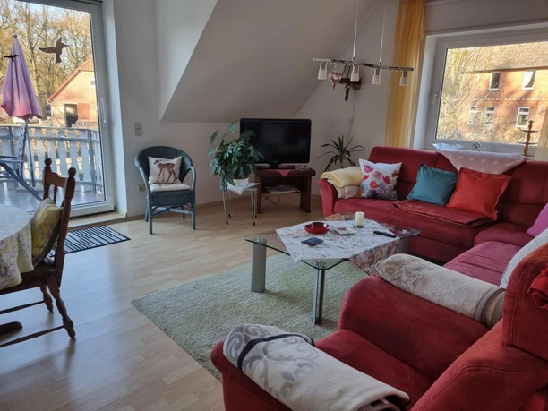 FeWo-Kühn-Wohnzimmer-4.JPG Wohnzimmer unter einer Dachschräge mit rotem Ecksofa, Glastisch, Fernseher und Balkontür.