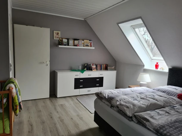 Schlafzimmer mit Doppelbett, Dachfenster, Kommode und Babybett auf Holzfußboden.