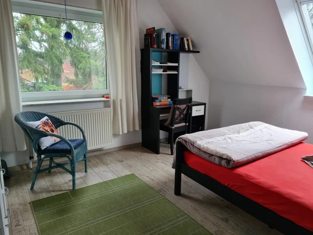 Helles Schlafzimmer mit Bett, Schreibtisch, Korbsessel am Fenster und Blick ins Grüne.
