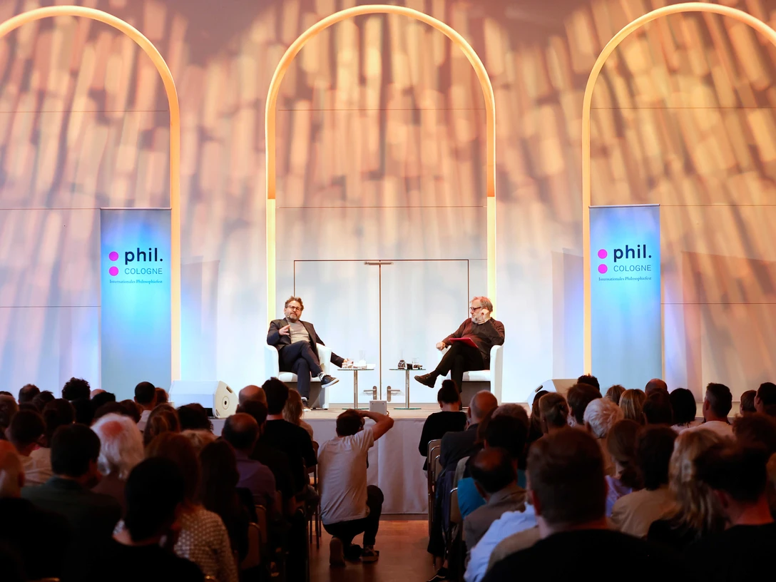 phil.Cologne Ein breites Publikum verfolgt eine Bühnenveranstaltung mit zwei Personen auf weißen Stühlen unter leuchtenden Bögen.A large audience watches a stage performance with two people on white chairs under illuminated arches.