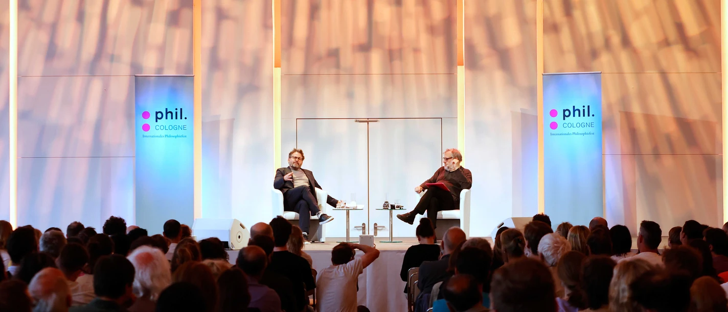 phil.Cologne Ein breites Publikum verfolgt eine Bühnenveranstaltung mit zwei Personen auf weißen Stühlen unter leuchtenden Bögen.