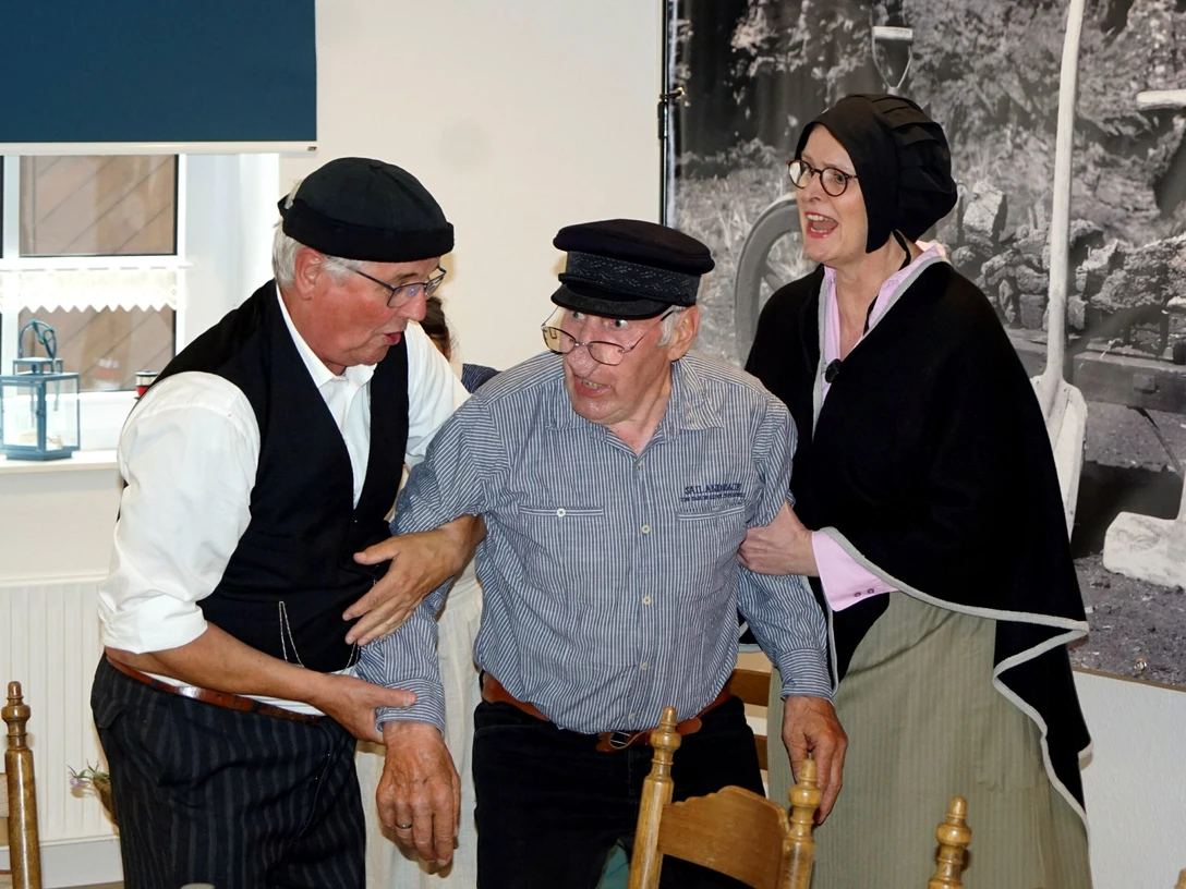 FehnTheater.jpg Drei Personen in historischer Kleidung, zwei stützen eine dritte Person, die sich an einem Stuhl festhält.