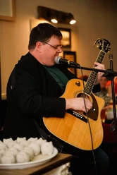 Christian Mettin.jpg Person sitzt und spielt akustische Gitarre vor Mikrofon in Innenraum mit unscharfem Hintergrund