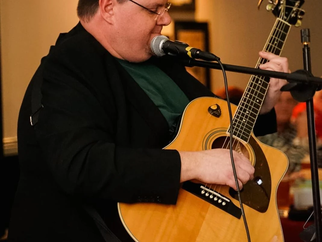 Christian Mettin.jpg Person sitzt und spielt akustische Gitarre vor Mikrofon in Innenraum mit unscharfem Hintergrund