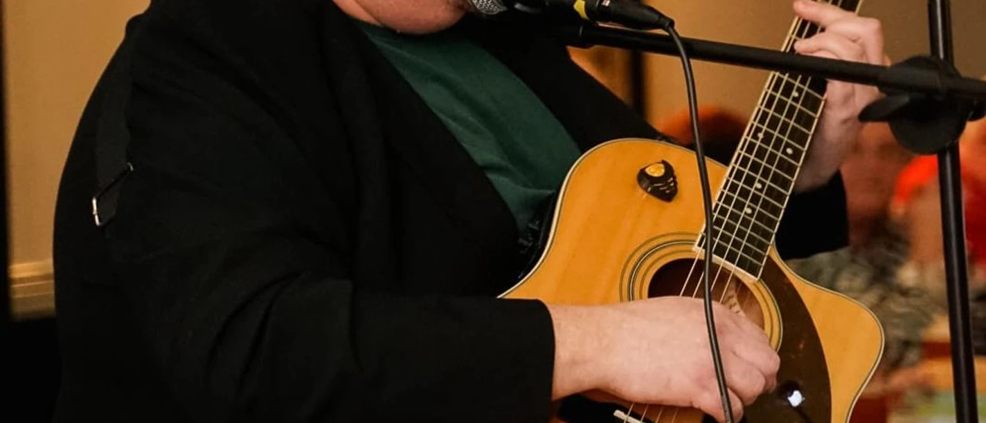 Christian Mettin.jpg Person sitzt und spielt akustische Gitarre vor Mikrofon in Innenraum mit unscharfem Hintergrund